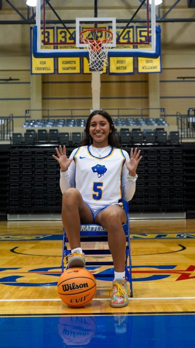 A young athlete in a basketball uniform sits on a chair, holding a basketball and smiling.